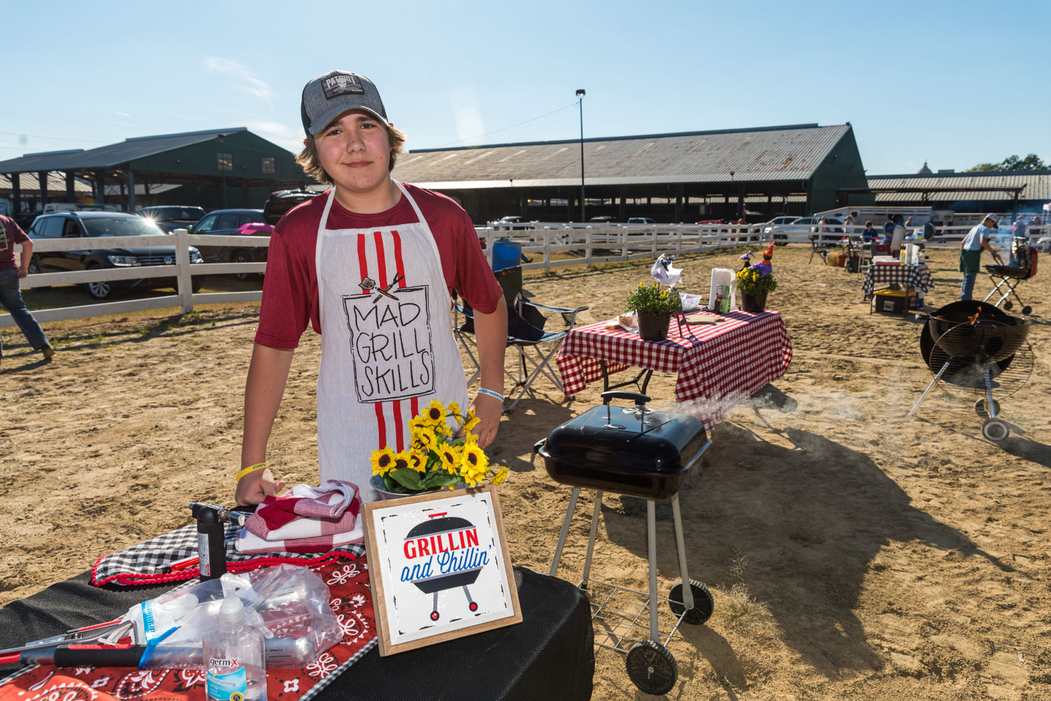 A boy, dressed in a baseball cap and apron, standing beside a smoking grill.