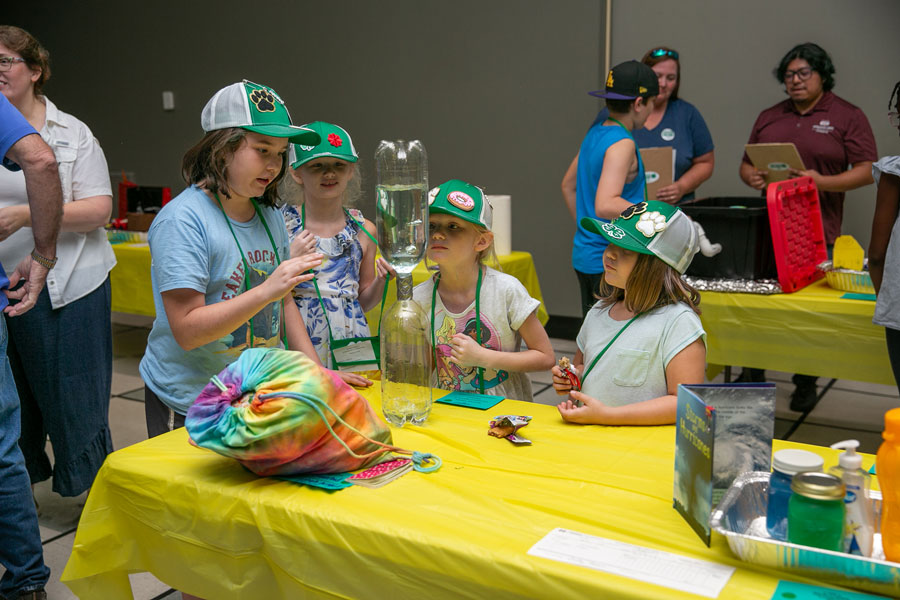 Four girls looking intently at two liter bottles joined at the mouth and containing liquid.