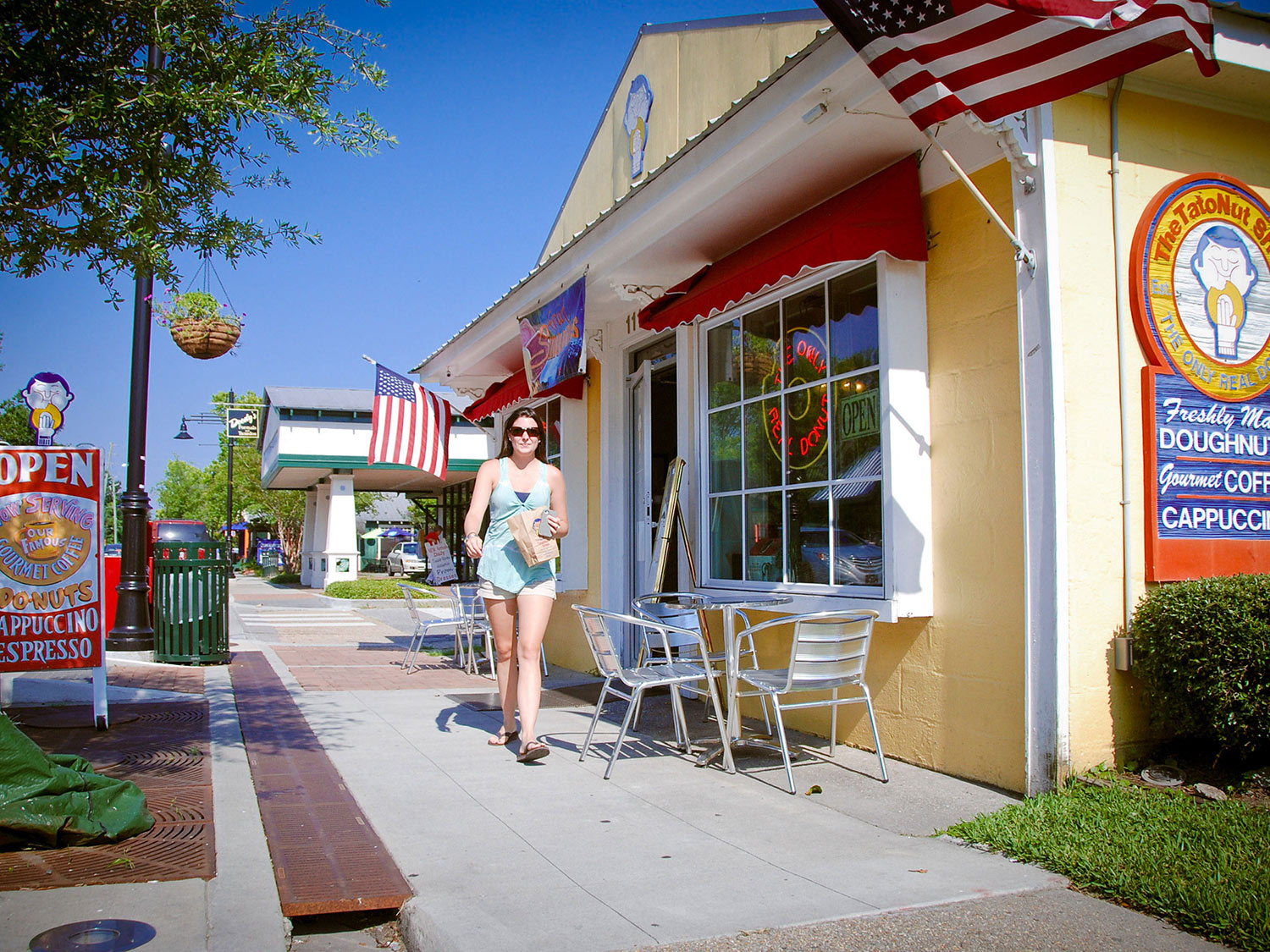 A woman holding a brown bag walks on a sidewalk in front of a yellow building with a sign that says, “Freshly Made Doughnuts.”