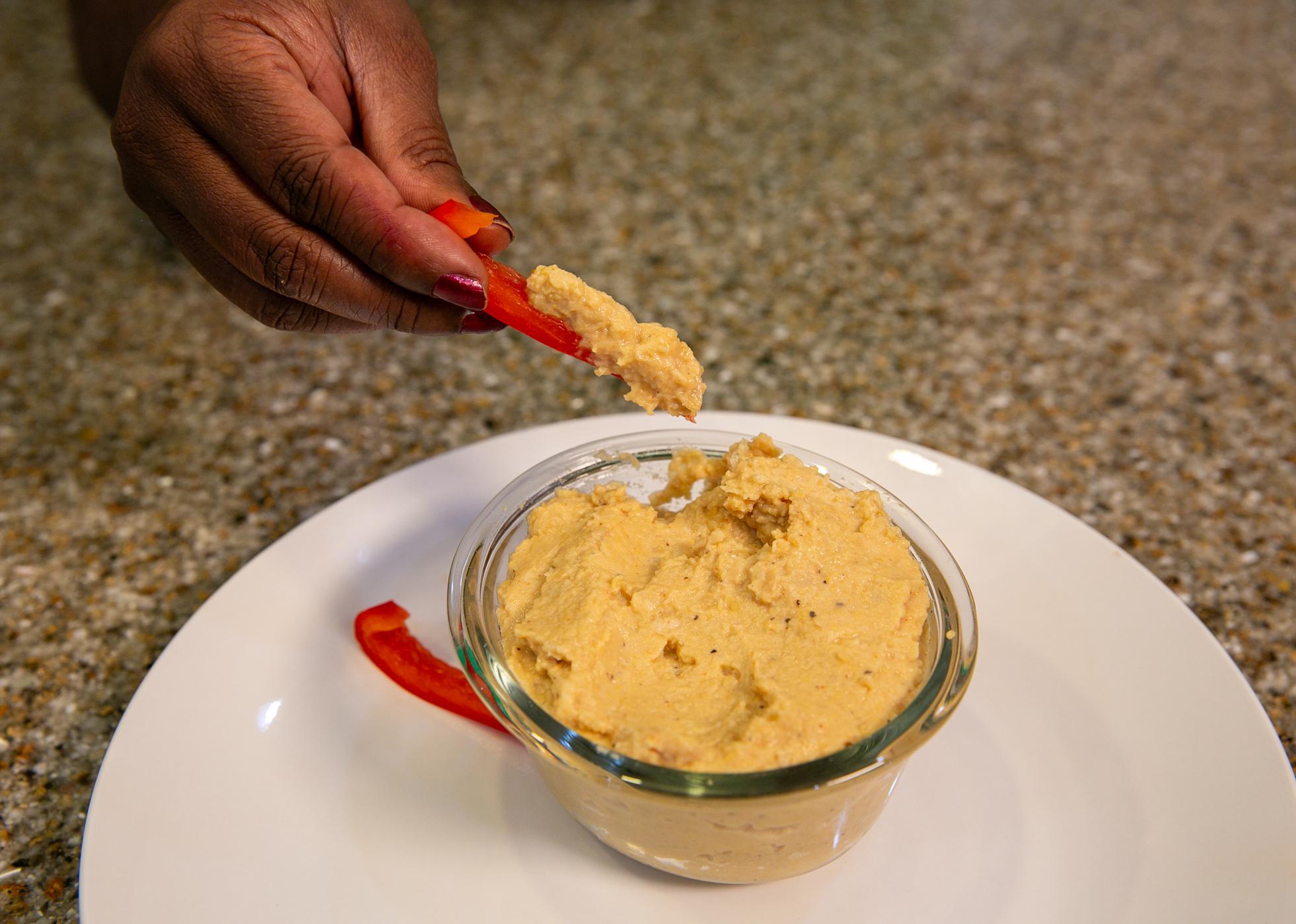 A hand is shown dipping a bell pepper strip into a bowl of garlic chickpea dip.