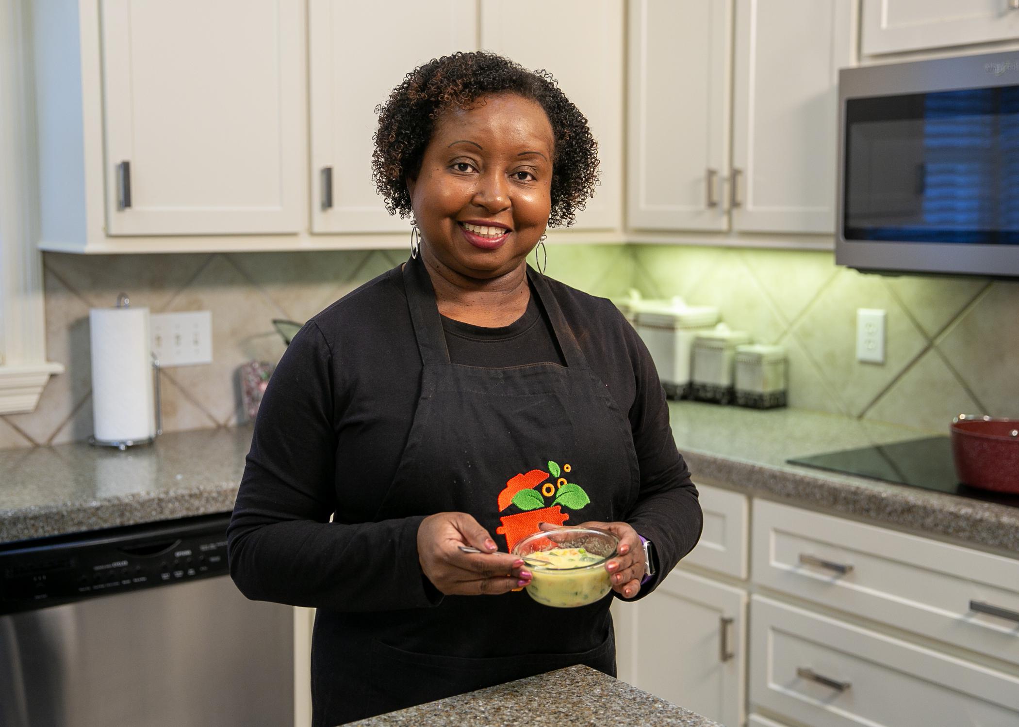 A woman stands in a kitchen and holds a bowl of soup.