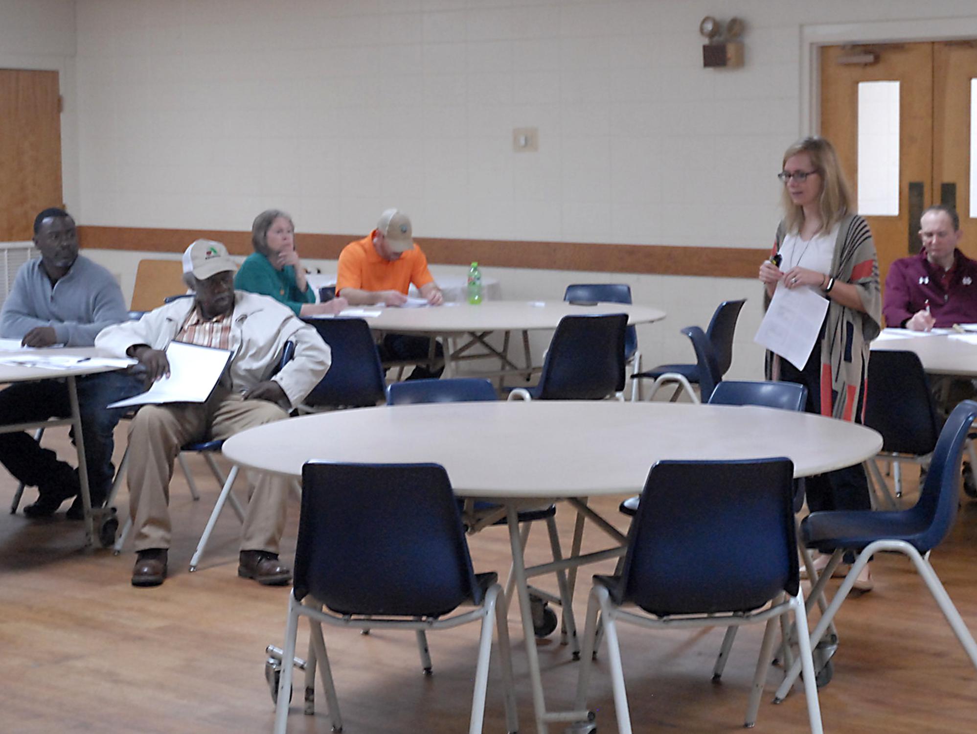 Blonde-haired woman with glasses in a large room speaking to a group of farmers.