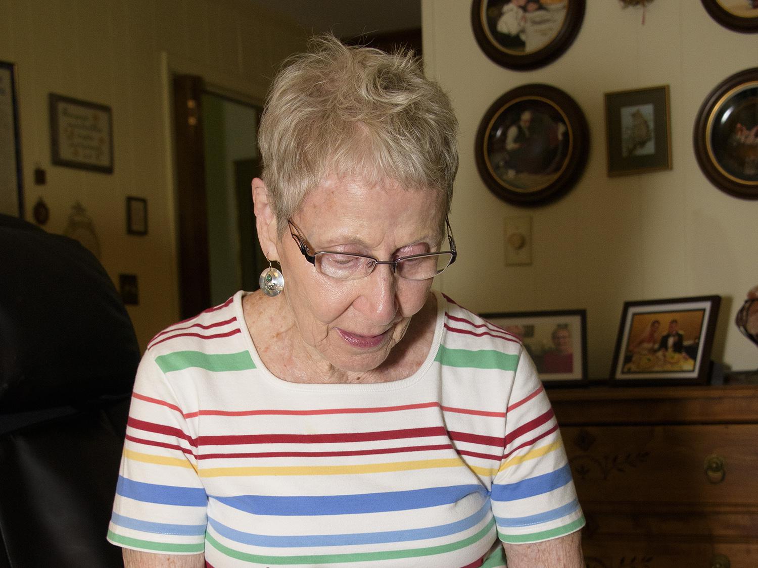 A gray-haired woman in blue shorts and a multi-colored striped shirt holds a pen over a pocket calendar to check appointments.