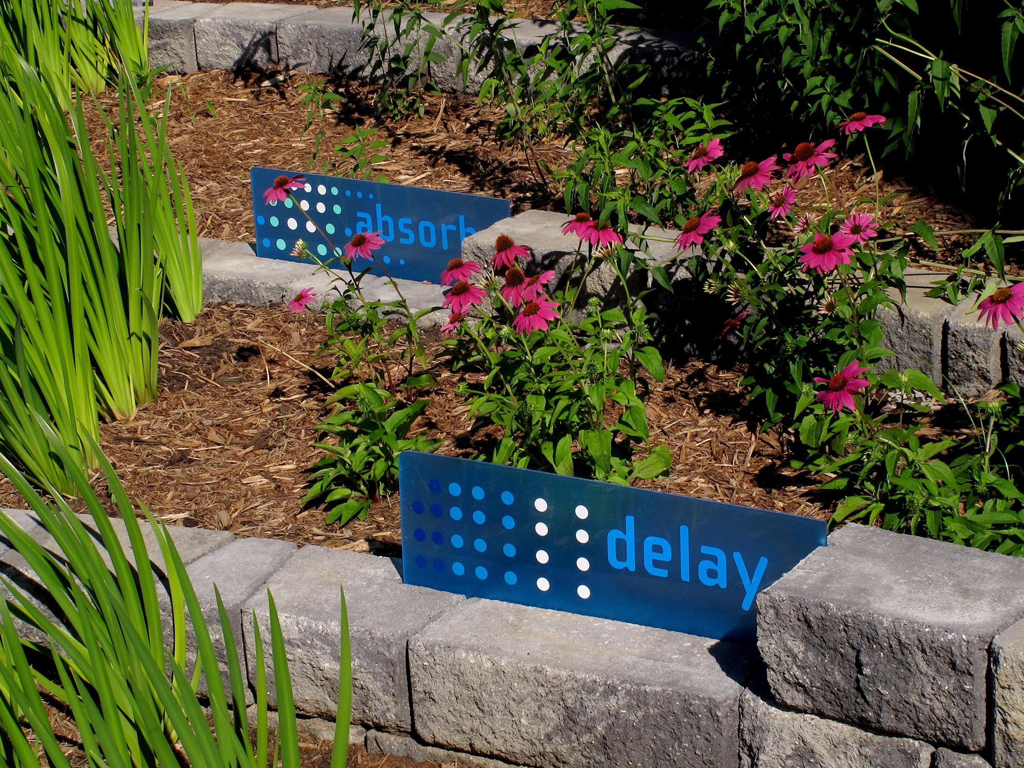 Blue signs stating “absorb” and “delay” mark brick tiers in a landscape growing pink flowers and reed-like stems.