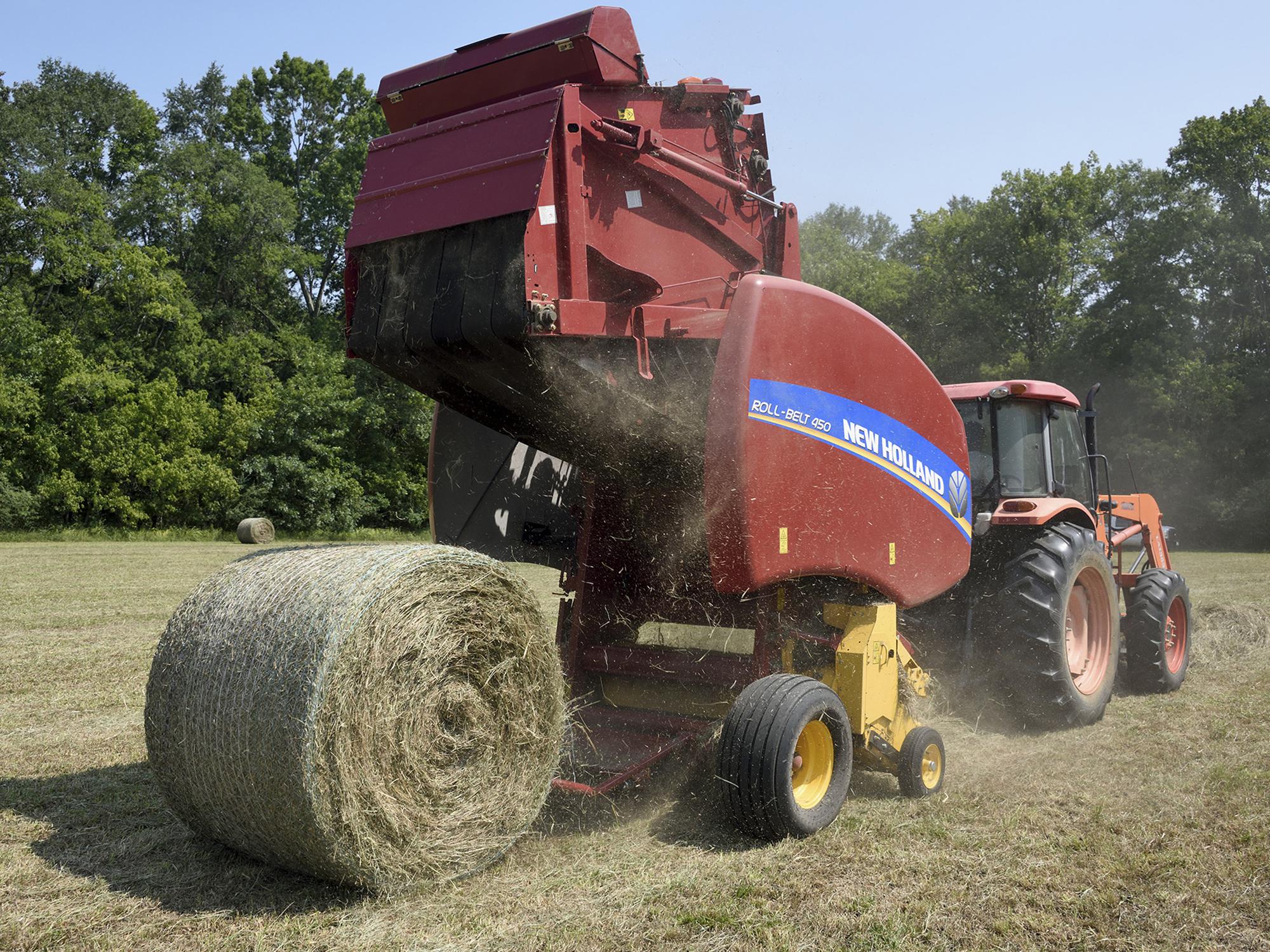 A red baler hitched to the back of an orange tractor drops a new, round bale of hay into a field.