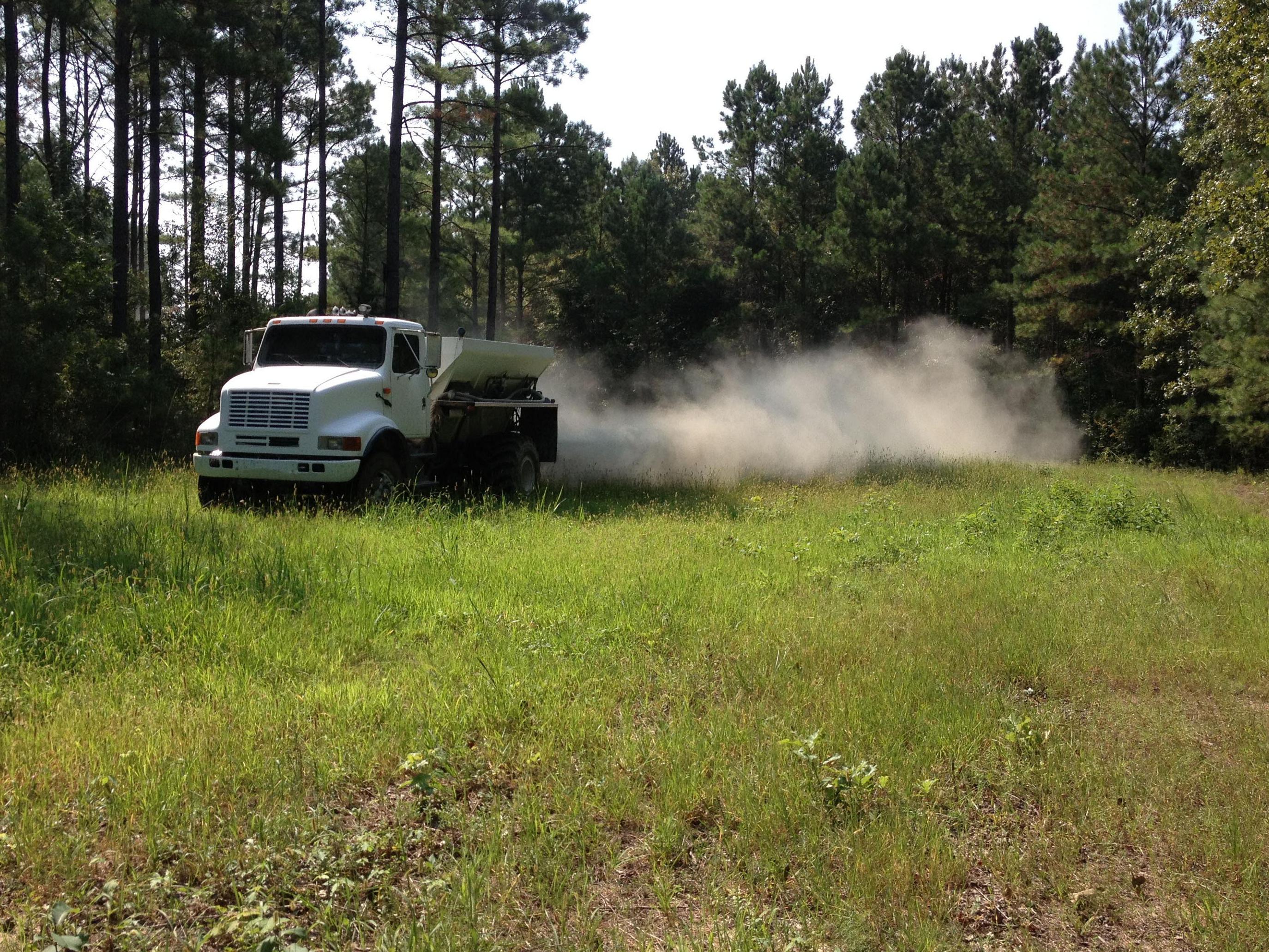 Dust billows out of a trailer on a large truck driving across a small, grassy area surrounded by tall trees.