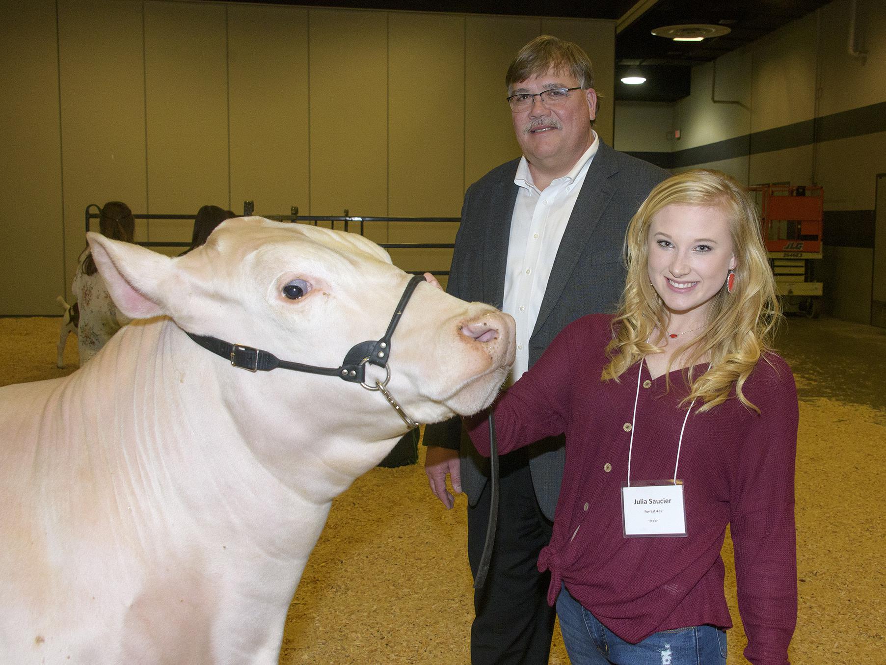 A teenage girl holds the halter on the face of her muscular, white steer as she and a tall man standing behind them look at the photographer.