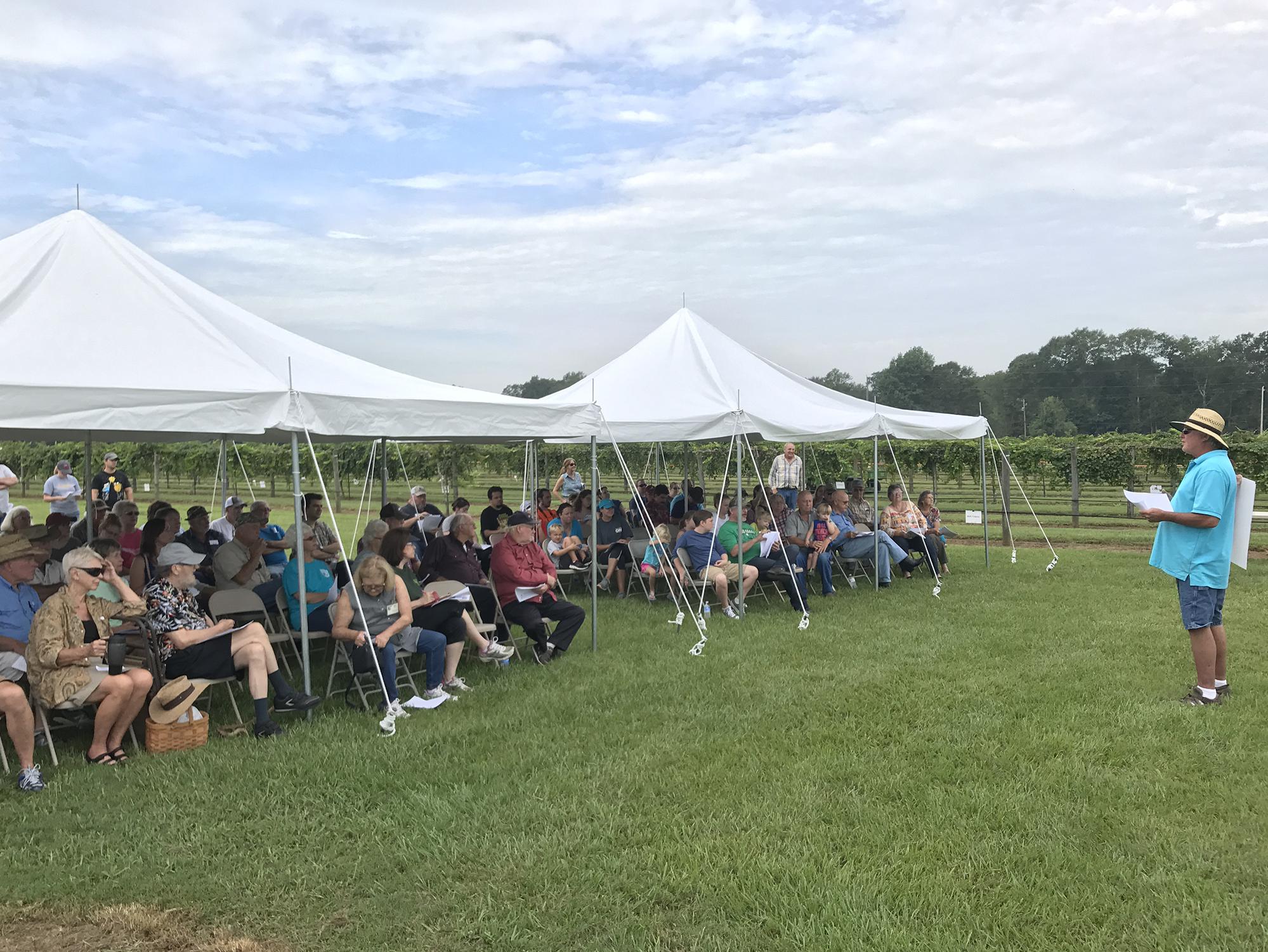 A crowd sits under tents as a speaker addresses them.