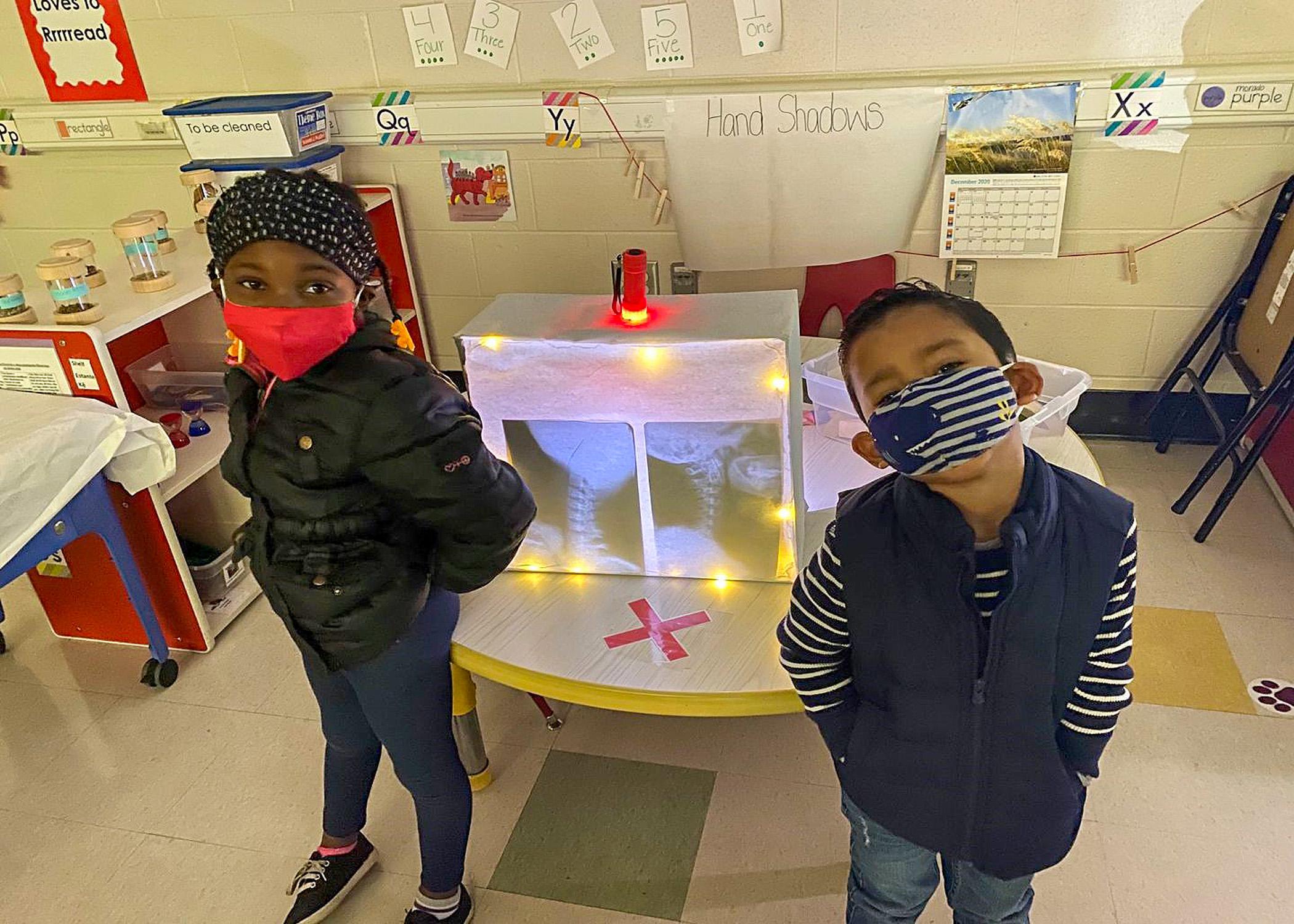 Two children in masks stand in front of an activity center in a classroom.