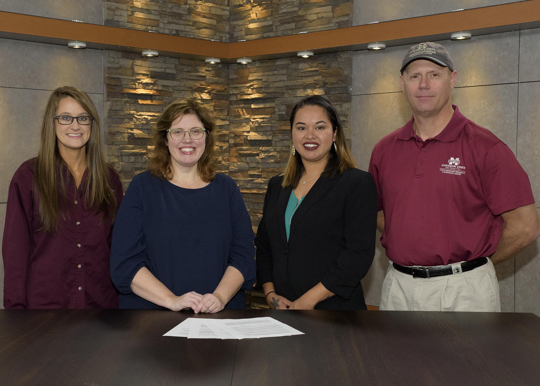 Three women and a man pose at a desk with a stack of papers.