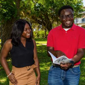 A young man pointing at a book he holds and woman, standing outside and laughing.