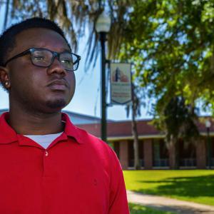 A young man, wearing glasses and a red shirt, standing outside and looking over the horizon.