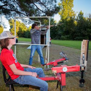 A boy with a gun at a shooting range with an older man watching him and a seated boy.