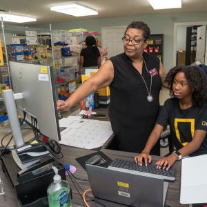 A woman points to a computer monitor as a teen girl looks with her hands on the keyboard.