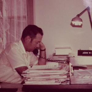 A man at a desk, surrounded by papers, and talking on the phone.