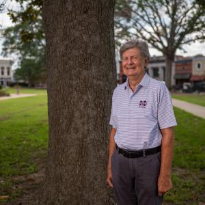 A man, wearing a Mississippi State University polo, smiling and standing in front of a tree.