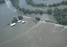 View from an airplane flying over extensive flood waters flowing over a levee and surrounding homes, farm buildings and crops.  