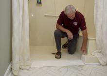 A man kneels in a walk-in shower, reaching down to a point in the entryway.