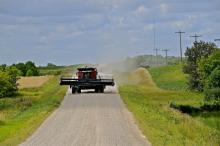 A red combine drives down a gravel road with farmland on both sides.