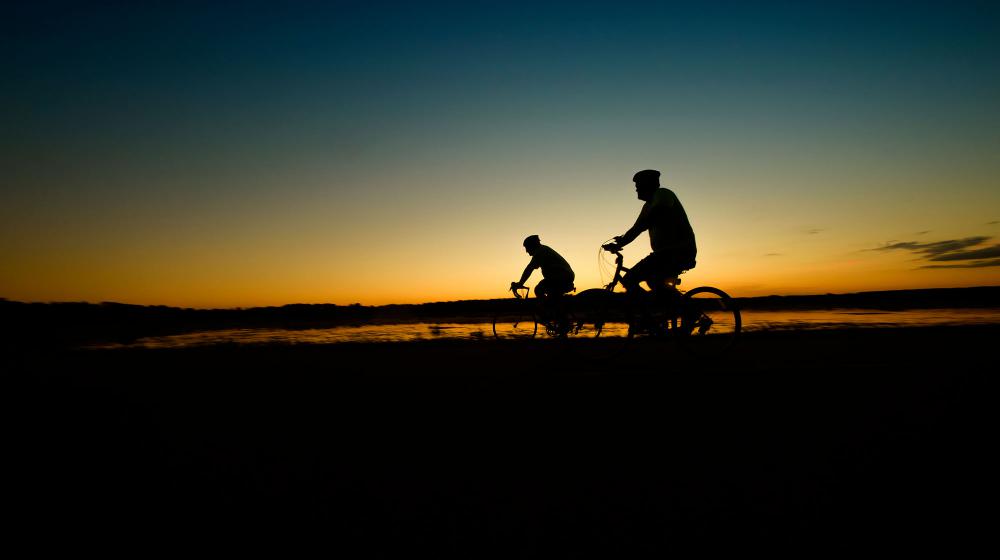 The silhouettes of two cyclists are seen in front of a blue and yellow sunrise that reflects on a lake below.