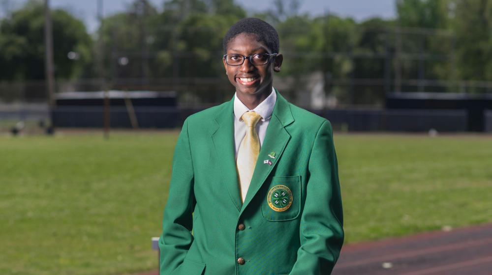 A young man wearing a green 4-H blazer stands smiling on a running track.