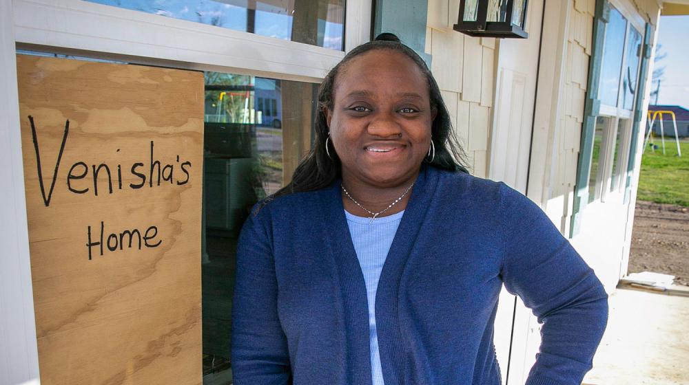 A woman standing beside the window of a house that has a sign with “Venisha’s Home” listed on it.