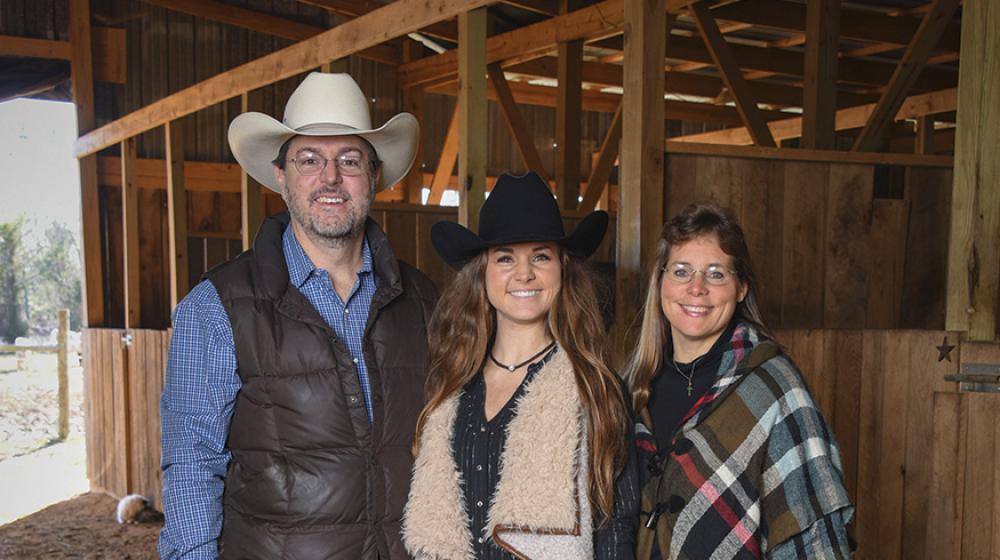 A teenage girl wearing a cowboy hat stand between her mom and dad, also wearing a cowboy hat, in a stable.