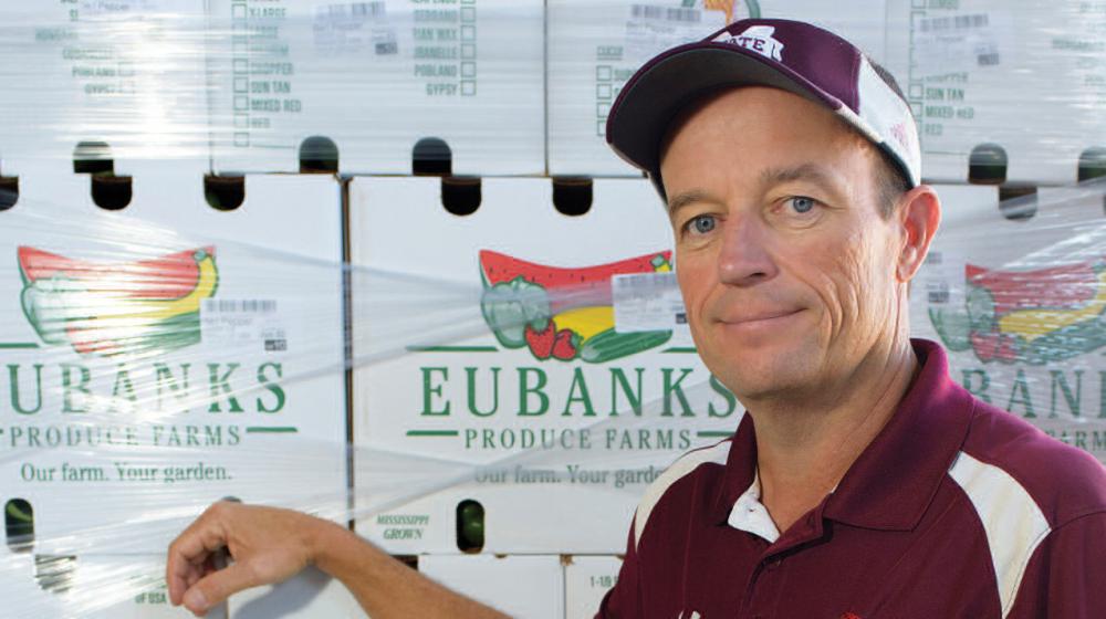 A man wearing a maroon shirt stands in front of a wall of boxes from Eubanks Produce Farm.