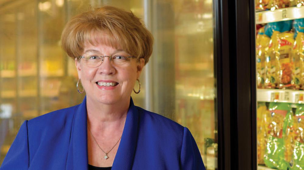 A smiling woman wearing a blue shirt stands in front of a frozen food aisle in a grocery store.