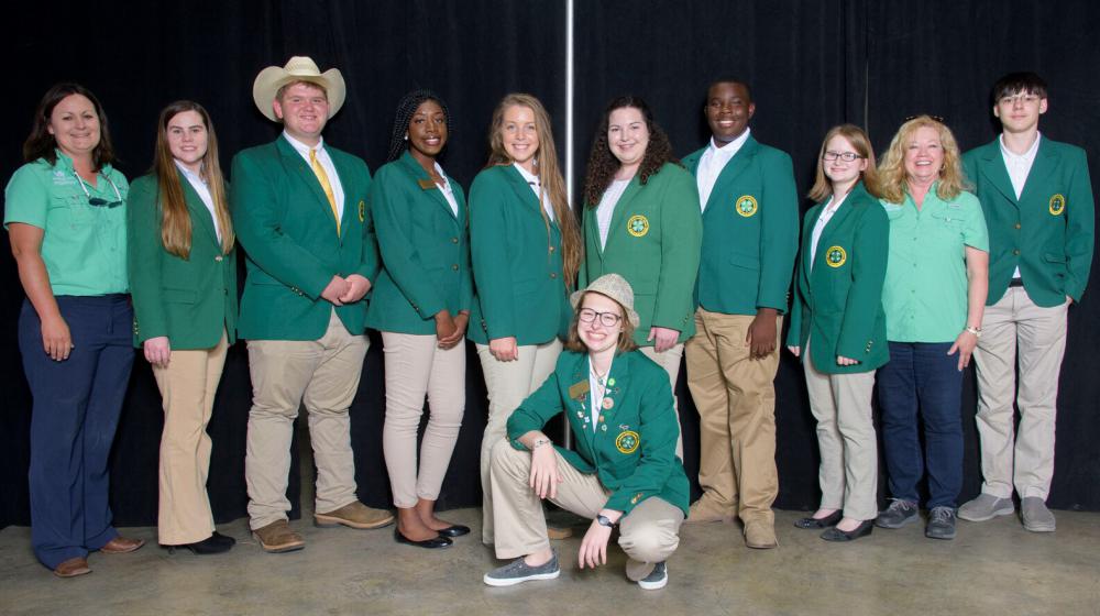 Eight teenagers wearing green blazers and two older women wearing green shirts stand smiling at the camera.