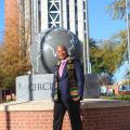 A man wearing a Jackson State University scarf and a navy suit jacket smiles in front of a sculpture on the JSU campus.