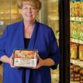 A smiling woman wearing a blue shirt stands in front of a frozen food aisle in a grocery store.