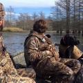 Two young men wearing camouflage sit in a small boat with a black dog, all looking out on the water on a sunny day.  