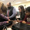 Two middle-school-aged girls and a middle-aged male teacher look on at another girl poking a hole in the side of a black tray for a science project.