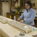 Small, cardboard containers in trays lie in a row on a workbench while a woman works with the contents of one.