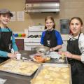 Three girls work with dough sheets at a table.