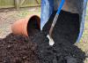 A shovel stands among soil from a wheelbarrow and a pot.