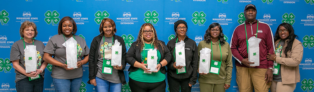 Seven women and one man hold up gift bags in front of a blue wall of MSU Extension and 4-H logos