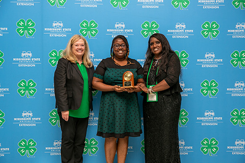 Three woman stand in front of a blue background with 4-H logo and MSU Extension logo repeated across